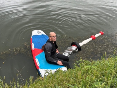 Mann in Neoprenanzug sitzt auf SUP-Board, daneben schwimmt ein Gerät mit roten Kugeln und Reifen
